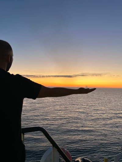 The scene is situated in the city of Unknown, Denmark, specifically at an unknown location at sea. The image depicts an adult male, viewed from behind and slightly to the right, standing on what appears to be the deck or structure of a vessel. The man, silhouetted against the bright horizon, has short, dark hair and wears a dark, short-sleeved t-shirt. His right arm is extended straight out, parallel to the horizon, with his hand open and palm facing upwards. This creates a forced perspective illusion where a large, distant offshore structure or vessel with a prominent central tower is aligned to appear resting on his open palm.
The sky above the horizon transitions from deep blue at the top to lighter blues, then to vibrant orange, yellow, and red hues near the horizon, indicating a sunset or sunrise. Several other smaller, indistinct vessel silhouettes are visible along the distant horizon line to both the left and right of the central offshore structure. The ocean surface is calm, dark blue, and exhibits subtle ripples, with some areas reflecting the warm tones of the sky. In the immediate foreground, a dark metal railing curves from the lower left towards the bottom center of the frame. Below the railing on the right, a white structure, likely part of the vessel's deck, is visible. A red and white object, partially visible in the bottom left, resembles a lifebuoy.