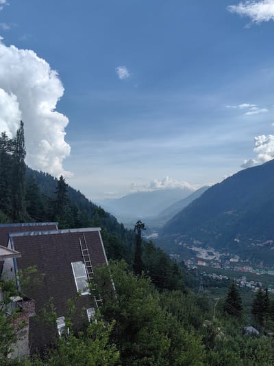The image depicts a scenic mountainous landscape with a village nestled in the valley below, likely in or near Siumsa, India. The foreground features the roof of a dark-shingled house with a large skylight and a ladder leaning against it. Dense green trees and foliage surround the house, partially obscuring it. In the mid-ground, a steep, forested mountain slope descends into a valley. The valley itself is dotted with buildings, roads, and a river or water channel. Several layers of mountains recede into the distance, fading into a hazy blue hue. The sky is a clear blue with scattered white clouds, one particularly large and puffy cloud mass dominating the upper left portion of the frame. Power lines and a transmission tower are visible on the forested slope. There are no people visible in the image.