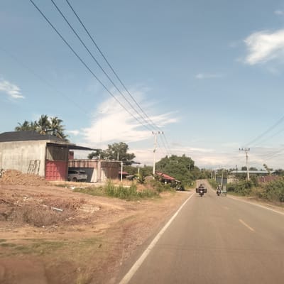 The image captures a daytime scene along a paved road in a rural or semi-rural setting. The sky is bright blue with scattered white clouds, and the sun appears to be high, suggesting mid-morning or afternoon.
In the foreground on the left, there is a large pile of reddish-brown dirt and dry, sparse vegetation. Behind this, a concrete building with a dark roof is under construction, featuring exposed concrete walls and red garage-style doors. A white car is visible, parked inside an open section of this structure. Palm trees and other green foliage rise above and around the building.
The asphalt road, which stretches into the distance, has a solid white line marking its right edge and a faded yellow dashed line down its center. Two motorcycles are traveling away from the viewer. The lead motorcycle has a rider wearing a helmet and a large rectangular box attached to its rear. Behind it, another motorcycle pulls a small, flat-bed trailer or sidecar, also with a rider.
Utility poles with multiple power lines line both sides of the road, extending into the background. Beyond the road and the vehicles, the landscape features more green trees and bushes, and various simple buildings or homes are interspersed amongst the greenery. No discernible text is visible in the image. The overall impression is one of everyday life and ongoing development in a tropical or subtropical region.