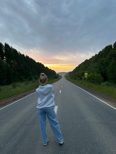 A person stands facing away from the camera, with their back to a straight, paved road that stretches into the distance. The road is bordered by dense, dark green trees on either side, with a faint glow of sunrise or sunset visible in the hazy sky at the vanishing point. The person is wearing a light gray hooded sweatshirt, light blue jeans, and athletic shoes. They have blonde hair pulled up in a bun. The sky above is filled with clouds in varying shades of gray and white, with a hint of pink and orange where the light is breaking through. A yellow traffic sign with a speed limit of 70 is visible on the right side of the road. The overall scene suggests an outdoor, rural setting, likely in or near Новая Казмаска, Russia, given the context.