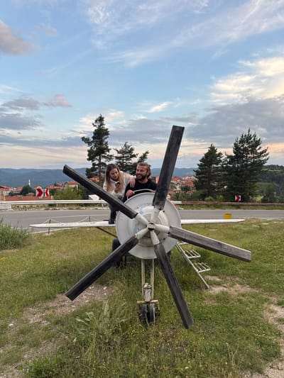 In the city of Sveta Petka, Bulgaria, a man and a woman are posing with a stationary propeller of a small aircraft. The propeller is mounted on a stand and appears to be made of wood and metal. The man, with blond hair pulled back, is seated and smiling. The woman, with long blonde hair, is standing behind him, looking at her phone and leaning on his shoulder. They are positioned in front of a rural landscape with rolling hills, trees, and houses in the background. The sky is partly cloudy with hues of blue, white, and a hint of sunset colors. The ground is covered in grass and a dirt path.
