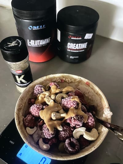 In Neubrandenburg, Germany, a close-up, top-down view captures a breakfast or post-workout meal preparation scene. Dominating the frame is a rustic, speckled bowl filled with frozen raspberries, whole cashews, and a drizzle of peanut butter. To the left of the bowl, a glass spice grinder with a black lid contains what appears to be cardamom, labeled "KARDEMOMME" and "TORG KRYDDERIER". In the background, two large black tubs of nutritional supplements stand prominently. The left tub, predominantly black with red and white accents, is labeled "L-GLUTAMINE HIGH BIOAVAILABLE CORE". The right tub, also black, is clearly marked with "CREATINE PURE MICRONIZED POWDER". The meal is positioned on a black digital scale, displaying a weight of "17" grams. The overall environment appears to be a kitchen counter.