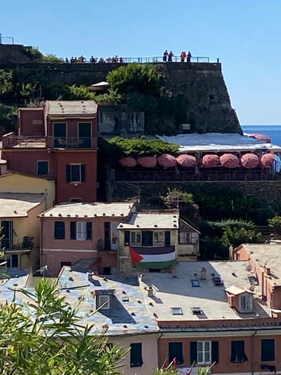 This image captures a multi-tiered view of Vernazza, Italy, under a clear blue sky.
In the upper section, a group of approximately 15-20 indistinct human figures is visible on a flat, open area atop a stone fortification or cliff edge. They appear to be standing and observing, positioned behind a light blue railing. To the right, the deep blue Tyrrhenian Sea is visible.
Below this fortification, and integrated into the hillside, is a restaurant terrace. This terrace features a row of eight circular patio umbrellas, all fully open and displaying a distinct red and white striped pattern. Below the umbrellas, dark tables and chairs are discernible, suggesting an outdoor dining area.
Further down the hillside, a cluster of traditional multi-story Italian buildings dominates the mid-ground. One prominent red-painted building features multiple windows with dark green shutters; its upper level includes two small balconies, one of which holds what appears to be a small table and two chairs. Adjacent and below are other residential structures in various hues, including pale pinks and yellows, with flat or gently sloping roofs of varied colors.
In the lower-mid ground, on the facade of a light beige two-story building, a flag is prominently displayed. The flag is rectangular, composed of three horizontal stripes—black (top), white (middle), and green (bottom)—with a red triangle on the hoist side that overlaps the black and white stripes. This flag is draped over a balcony or window railing.
The foreground contains sparse green foliage, partially obscuring the lowest visible buildings. Sunlight illuminates the scene, creating distinct shadows on the roofs and buildings.