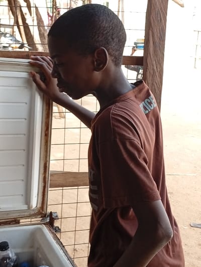 A young Black person, appearing to be a child or teenager, is captured in profile looking into an open chest freezer in Fanisau, Nigeria. They are wearing a plain brown short-sleeved t-shirt, on the back of which the word "POLICE" is partially visible in light-colored text. Their right hand rests on the edge of the freezer opening.
Inside the freezer, several bottles are visible, some containing dark liquids. The freezer's edges show signs of wear and some rust. The scene appears to be an outdoor or semi-outdoor vendor stall or shop, characterized by a metal grid fence or wall in the background and sturdy wooden posts. The ground beneath is light-colored, possibly sand or packed earth.
The bright, natural lighting indicates it is daytime, likely a clear and warm day. The person's expression is neutral and focused on the contents of the freezer, suggesting they are either choosing an item or inspecting the inventory. The image provides a glimpse into a moment of daily activity in this local Nigerian setting.