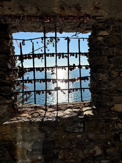 A view from within a weathered stone structure in Monterosso al Mare, Italy, looks out towards the Mediterranean Sea. The foreground features an irregular window opening framed by coarse, dark, shadowed stone blocks. A dark, metallic grate, composed of vertical and horizontal bars, spans the entire aperture. Hundreds of small, colored padlocks, commonly referred to as "love locks," are affixed to the horizontal and vertical bars of this grate, densely covering its surface.
Beyond the grate, the sea is visible under a clear, bright blue sky. A prominent, intensely bright sun glare reflects on the water's surface, creating a wide, shimmering column of light that extends from the distant horizon towards the foreground. Parts of dried plant matter, including thin stems and spherical seed pods, are entangled within the upper left section of the metal grate. A portion of a green, multi-lobed palm frond is visible through the left side of the window opening, suggesting coastal vegetation just outside. No individuals are present in the scene. The lighting indicates direct sunlight outside, contrasting with the heavily shadowed interior of the stone structure.