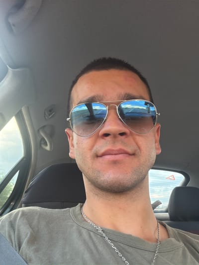 This media file shows a close-up, low-angle selfie of a person from the chest up, taken inside a vehicle. The subject is wearing aviator sunglasses with reflective blue lenses, a grey t-shirt, and a silver chain necklace. The sunglasses reflect a scene of a road with a blue sky and some clouds, suggesting they are traveling. The background includes parts of the car's interior, such as the ceiling, a headrest, and a window with a triangular warning sign. The lighting suggests it is daytime, possibly late afternoon, with sunlight streaming through the window. The person's facial expression is neutral, and the overall mood is casual. The image appears to be taken in Mirovo, Bulgaria.