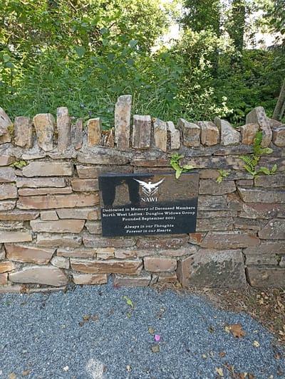 A black granite plaque is affixed to a rough stone wall in Dungloe, Ireland, with a white bird emblem at the top. The inscription on the plaque reads: "NAWI Dedicated in Memory of Deceased Members North West Ladies - Dungloe Widows Group Founded September 2001 Always in our Thoughts Forever in our Hearts." The wall is constructed from irregular, weathered stones, some of which are partially covered in moss. Behind the wall, lush green foliage and trees create a natural backdrop. The ground in front of the wall is covered with gray gravel and scattered dry leaves.