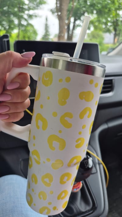 This media file shows a person's hand holding a white tumbler with a yellow leopard print pattern. The tumbler has a silver rim and a white handle. A light-colored straw is inserted into the tumbler's lid. The background suggests an indoor setting, possibly a car, with a dark dashboard, a gear shift, and a yellow cable visible. Through a window, lush green trees and foliage can be seen, indicating an outdoor scene with daylight, likely in the late spring or summer. The person's fingernails are painted a light pink. The overall impression is casual and everyday. There is no visible text.