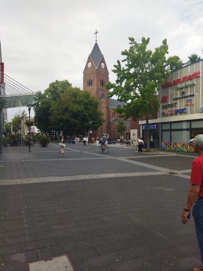 The image depicts a street scene in Langenfeld (Rheinland), Germany. In the background, a brick church tower with a clock is prominent. Adjacent to the church, a modern building with shops is visible, featuring signs for "H&M," "DEPOT," "INTERSPORT," "dm," and "TOM TAILOR." The street is paved with dark gray bricks and a lighter gray border. Several people are present, including a woman with a shopping bag standing in the foreground, a man cycling in the middle ground, and a group of people gathered near the shops. Trees and outdoor seating areas for a cafe are also visible. The sky is overcast. A man wearing a red shirt and blue jeans is partially visible in the lower right corner of the image, holding a blue object in his right hand.
