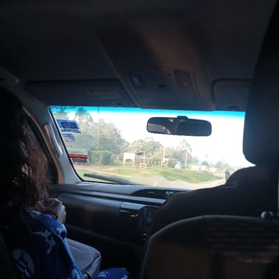 The image captures a view from within a vehicle, looking forward through the windshield onto an unpaved road in Butsotso central ward, Kenya, during daytime.
**People:**
- A person with long, dark, wavy hair occupies the front passenger seat, facing forward. They are dressed in a blue garment with a white circular pattern and hold a light-colored handbag.
- The rear of a second person's head and right shoulder, wearing dark attire, is visible in the driver's seat on the right.
- A lone individual in dark clothing is walking on the distant right side of the unpaved road.
**Vehicle Interior:**
- The vehicle's dashboard and interior trim are dark-colored.
- The windshield displays two blue-and-white stickers and one red-and-white sticker affixed to its upper-left section.
- A rearview mirror is centrally mounted above the dashboard.
- An overhead console with various buttons is visible on the ceiling.
- A single windshield wiper blade rests horizontally on the lower portion of the windshield.
**Exterior Environment:**
- The vehicle is situated on a light-colored, unpaved road.
- The surrounding landscape is characterized by abundant green vegetation, including numerous trees and bushes.
- On the left side of the road, a simple, open-sided structure with a light-colored roof and visible support posts is present.
- Further along the road on the right, several distant, low-rise structures are discernible.
- The sky is bright and clear, suggesting daylight conditions.
