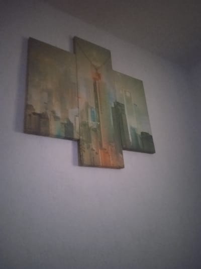 An abstract canvas art piece depicting a city skyline is mounted on a textured, off-white wall. The artwork is composed of four rectangular panels arranged in a staggered, cross-like formation. The dominant colors are muted greens, grays, and some orange/brown hues, suggesting a hazy or artistic interpretation of urban structures. The perspective is from below, looking up at the artwork. The scene is indoors, with the wall being the only visible element of the environment. There are no people, animals, or discernible activities. The lighting appears to be ambient, casting a slight shadow on the wall to the left of the artwork. There is no information provided about the location in Nihoreni, Moldova, and therefore, it cannot be included in the description.