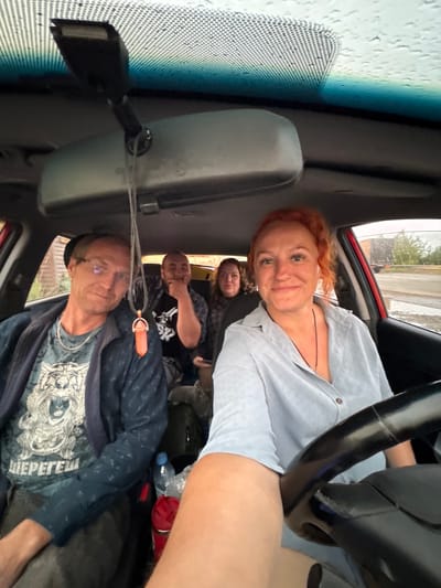 A selfie taken from inside a car shows four people. The driver, a woman with reddish-orange hair styled in a high ponytail, smiles at the camera. She is wearing a light blue collared shirt and has a necklace with a pendant. To her left, a man wearing glasses and a dark blue jacket over a graphic t-shirt, is looking towards the camera with a slight smile. Behind them, two other individuals are visible. The passenger behind the driver is partially obscured, but appears to be a woman. The passenger behind the driver in the front seat is a man with a beard, looking towards the left. The car interior is visible, including the rearview mirror, the ceiling lining, and the steering wheel. Outside the car, through the windows, a street scene with trees and a road can be seen, suggesting an outdoor environment. The sky appears overcast with some visible raindrops on the windshield. The location is identified as Sovkhoznyi, Russia, which is a settlement in the Sverdlovsk Oblast.