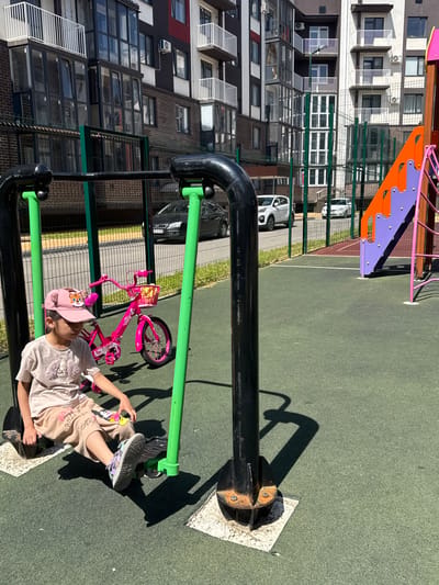 A young girl is sitting on a playground exercise machine in a city park in Vyazniki, Russia. She is wearing a light-colored t-shirt, light brown shorts with cartoon characters, and a pink baseball cap with a tiger design. Her feet are on the machine's footrests, and her hands are resting on the handles. To her right, a pink children's bicycle with a basket is parked. Behind the playground equipment, a fence separates the park from a residential building with multiple balconies and windows. Several cars are parked in front of the building. The ground is covered with green rubberized material, and the sky is clear and blue.