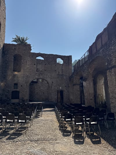 The image shows an outdoor area in Dolceacqua, Italy, featuring rows of empty black plastic chairs arranged on a cobbled ground. Behind the chairs, the remains of ancient stone structures are visible, with arches, openings, and crumbling walls. The ruins suggest a historical or archaeological site, possibly a former amphitheater or performance space, given the seating arrangement. Sunlight casts shadows from the chairs onto the ground, and a bright sun is visible in the upper right portion of the sky. The architecture and natural elements indicate a warm, sunny day in an old European town.