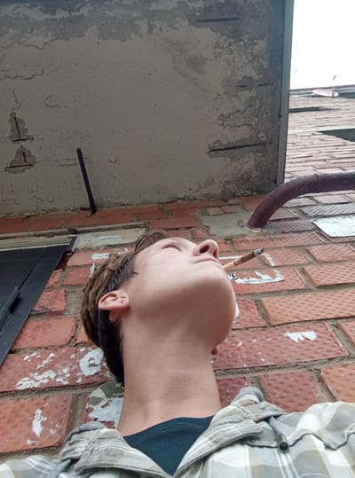 A low-angle medium shot captures a person smoking a cigarette outdoors against a brick wall and a concrete structure. The person's head is tilted back, and their eyes are closed, with smoke exhaling from their mouth. They are wearing a plaid shirt over a dark t-shirt. The background consists of a rough, weathered concrete overhang with exposed rebar, a red brick wall with remnants of posters, and a large, dark metal door on the left. A thick, dark pipe curves into the scene from the right. The overall environment suggests an urban or semi-urban setting. No specific location in Gdov, Russia, can be determined from the visual information.