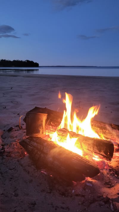 A campfire burns on a sandy beach at dusk in Zaostrov'e, Russia. The fire is situated on the sand, with logs smoldering and flames reaching upwards. A metal mug with a handle is placed next to the campfire. In the background, a calm body of water stretches to the horizon, with a dark treeline visible on the far shore. The sky is a deep blue, indicative of twilight, with a few wispy clouds. The scene suggests a peaceful, outdoor setting.