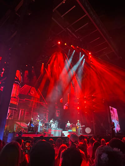 This image captures a live music concert at the Les Schwab Amphitheater in Bend, Oregon, United States. The scene is illuminated by intense red stage lighting, with several distinct white light beams penetrating atmospheric haze. The stage features a large LED screen backdrop displaying a detailed, stylized rendering of a distressed house-like structure in red and dark tones. Approximately six musicians are visible on stage, performing. A central figure, wearing a hat, plays a guitar. A drummer is positioned behind a drum kit on a raised platform, with other musicians operating various instruments including guitars and keyboards. The foreground is occupied by a dense crowd of concert attendees, viewed from behind, with some individuals holding mobile phones vertically, recording the event. The overhead structure of the amphitheater, including a partial sign indicating "T HOMES AMPHITHEATER," is visible in the upper portion of the frame.