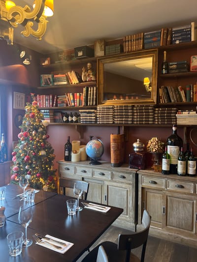 This image depicts the interior of a dining establishment located in Kortrijk, Belgium, presenting a corner of a room adorned with festive and decorative elements. The scene is illuminated by warm ambient light from a chandelier, wall-mounted spotlights, and a decorated Christmas tree.
In the foreground and extending to the midground on the left, a dark rectangular dining table is set with multiple clear drinking glasses and cutlery resting on white napkins or placemats. Several dark wooden chairs with light-colored seats are positioned around the table. To the left of the table, a fully decorated Christmas tree stands prominently, illuminated by numerous warm lights and adorned with red, white, and silver baubles and ornaments. A bottle of sparkling wine is visible on a cabinet just to the left of the Christmas tree.
The background wall is painted a reddish-brown color and features multiple dark wooden shelves stacked with numerous books of varying sizes and bindings. On the upper left shelf, a framed photograph of two individuals and a bust of Jesus Christ with a raised hand are visible. Centered on the wall above eye level is a large rectangular mirror with an ornate gold frame, reflecting an interior light fixture. Below the mirror, a long row of horizontally stacked books creates a decorative frieze. Further right on the shelves, a bottle of champagne and additional books are arranged.
Below the shelves, two distressed light-wood sideboards with closed doors and drawers run along the wall. The left sideboard displays a dark wine bottle, three white pillar candles (one glowing orange), a small blue globe on a stand, and a large object resembling an old, oversized book or scroll container. The right sideboard holds a dark glass jar with a metallic lid, an arrangement of dried decorative items such as acorns, an oversized wine bottle labeled "MUFFINS" (partially legible), and four standard-sized wine bottles, one with a green label. The floor appears to be dark wood or laminate. No individuals are present in the scene.