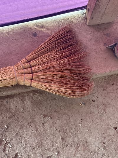 The image shows a close-up view of a broom made of dry straw, lying on a cracked, dusty, concrete surface. The broom is bound with multiple thin, light-colored ties. In the background, a section of what appears to be a wooden beam is visible, and above that, a surface with a purple hue, possibly a piece of fabric or plastic sheeting. A portion of a red and silver object, possibly a tool or equipment, is also visible in the upper right corner. The scene appears to be outdoors, possibly in a construction or storage area. There are no people or animals visible in the image. The lighting suggests it is daytime. The overall impression is one of a rustic, possibly abandoned or utilitarian setting. There is no identifiable information within the image that would allow for the determination of its specific location within the city of International, Kyrgyzstan.