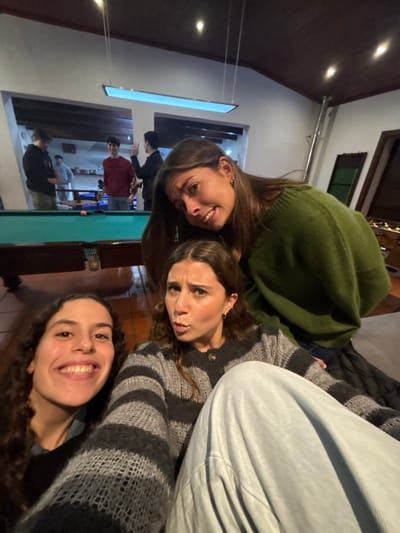 Three women are angled from the bottom right to the top left of the image, posing closely together. The woman at the top left is wearing a green sweater and smiling. Below her is a woman in a striped sweater with a neutral expression. The woman at the bottom right has curly hair and is smiling broadly. Behind them is a billiards table, and a group of people can be seen playing billiards in the background. The setting appears to be a casual indoor space with visible lighting fixtures.