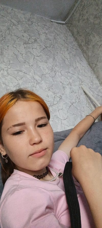 A young woman with orange-tinted hair and light skin is pictured in a close-up, angled shot. She is wearing a pink t-shirt, a black choker, and a pearl necklace with a heart-shaped pendant. She has prominent eyeliner and thin eyebrows. Her left arm is extended, and her right arm is bent, with her hand near her face. The background is a wall with a grey, marbled pattern and a white pipe or tube. The scene appears to be indoors, possibly in a residential setting. The location within Александровский Завод, Russia, is not determinable from the image.