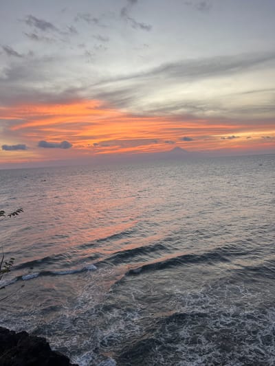 The image presents a wide, elevated view of a sunset over the ocean in Desa Batu Layar Barat, Indonesia. The sky occupies the upper portion, displaying a gradient from light grey and white streaky clouds at the top, transitioning into vibrant bands of orange, red, and yellow stratiform clouds across the mid-horizon. The vast expanse of the ocean fills the lower two-thirds of the frame, its surface reflecting the sunset's colors with pinkish-orange hues blending into deep grey-blue. Distinct waves with white foam are visible in the foreground, rolling towards the observer. On the distant horizon, two small, dark, elongated vessels are discernible on the water. A prominent, dark, triangular-shaped mountain or volcanic peak is silhouetted against the colorful sky on the right side of the distant horizon. The immediate bottom-left foreground reveals a dark, rocky cliff edge with sparse, dark foliage, indicating the elevated viewpoint. No individuals or specific human activities are directly visible.