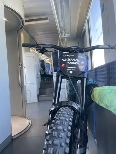 A first-person perspective shot from inside a train carriage shows a mountain bike parked vertically. The bicycle's handlebars are in the foreground, followed by the front fork and tire. A black and blue number plate is attached to the handlebars, displaying "CANYON ENDER" and several sponsor logos, including SRAM and FOX. The train carriage has blue and gray seats visible in the background, along with a window. The floor is gray, and to the left, a curved doorway with a silver handle is visible. The environment suggests the bicycle is being transported by train, possibly to or from Merichleri, Bulgaria, given the context. There are no people clearly visible in the frame, only the presence of the bicycle and the interior of the train.