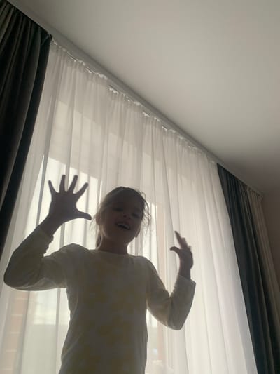In a low-angle shot, a young girl with blonde hair and fair skin is captured from below, leaning towards the camera. She is wearing a light-colored long-sleeved shirt with a subtle yellow floral pattern. Her mouth is open in a wide smile, and her eyes are bright, conveying a sense of excitement or playfulness. Her right hand is outstretched and open, fingers spread, creating a shadow on the sheer white curtains behind her. The curtains are gathered and pleated, extending across the frame and illuminated by a bright light source, likely daylight filtering through a window.

Darker, thicker curtains are visible at the bottom and top of the image, suggesting a dual-layer curtain system. The overall setting appears to be indoors, possibly a bedroom or living room, with the natural light creating a slightly ethereal and dramatic effect. The perspective from below emphasizes the girl's playful posture and the texture of the curtains. There is no visible text in the image. The image seems to capture a spontaneous moment of childlike joy and mischief.