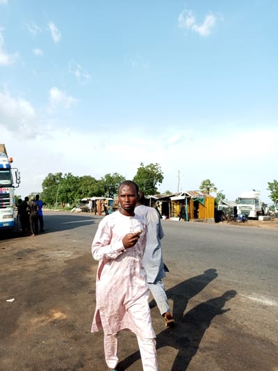 The image depicts a scene in Gadao, Nigeria, under a bright, partly cloudy sky, suggesting it is daytime, likely mid-morning or afternoon. The primary subjects are several men, most of whom are dressed in traditional Nigerian attire. In the foreground, a young man wearing a light pink patterned traditional outfit, possibly a 'babanriga' or 'agbada', is walking and looking directly at the camera. He holds a small object, perhaps a phone or a wrapper, in his right hand. Behind him, another person in a light blue traditional outfit is also walking, their face not fully visible.

To the left of the frame, a white and blue truck with "LAGOS" visible on its side is parked, with a group of men gathered near it. Further in the background, there are several market stalls or roadside shops with corrugated iron roofs and open fronts, where goods are likely being sold. Trees with lush green foliage are scattered throughout the scene, indicating a possibly tropical or sub-tropical climate. Another white truck is visible in the middle ground to the right, along with smaller vehicles and more market structures.

The overall atmosphere appears to be that of a busy roadside market or a truck stop. The light suggests a sunny day, and the clear sky with scattered clouds points to fair weather. The individuals in the image do not display overt emotions, appearing focused on their activities or posing for the camera. The presence of trucks and market activities suggests a transportation hub or commercial area.
