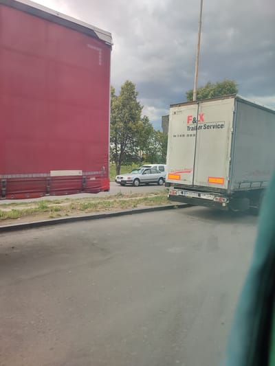 A street scene in Barmaky, Ukraine, features a red truck trailer on the left, partially obscuring a lighter-colored trailer with the text "F&X Trailer Service" prominently displayed. The lighter trailer has a website address "www.Stressfrei-Mieten.de" and a phone number "0 18 05-28 95 55" visible. A silver sedan is parked behind the lighter trailer. A utility pole stands tall behind the trailers, with trees and a cloudy sky in the background, suggesting overcast weather, possibly late afternoon. The road surface is visible in the foreground, with a curb and grassy verge. The license plate on the rear of the lighter trailer reads "AM 2095 XO UA UA". The scene appears to be a roadside service area or a busy street with parked vehicles. There are no people visible in the image.