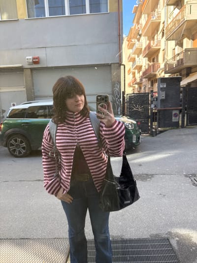 A female individual stands on a city street in Arezzo, Italy, capturing a mirror selfie with a smartphone held in her right hand. She has shoulder-length brown hair with bangs and wears a light pink and dark red striped zip-up jacket over a dark top, dark blue jeans, and a dark belt. A gray backpack is on her shoulders, and a black shoulder bag hangs from her left side. Multiple rings are visible on her right hand. The street surface is paved, with a metal grate in the immediate foreground and some wet patches on the ground. To the left, a light-colored building with white-framed windows and a garage door is visible, in front of which a dark green Mini Cooper Countryman is parked. Two red and white fixtures are mounted on this building. To the right, a sequence of cream-colored multi-story buildings with balconies extends down the street under a clear blue sky. A dark metal gated entrance with a parking barrier system and a sun-like emblem is also visible to the right. The scene is illuminated by direct sunlight, casting some shadows.