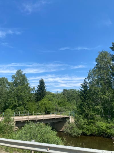The image depicts a bridge over a body of water, surrounded by lush green foliage and trees under a bright blue sky with scattered white clouds. The bridge appears to be constructed of concrete and metal, with a railing along its length. The water below is dark and reflects the surrounding greenery. The scene is set in a natural, possibly rural, environment. While the location is stated to be in Bolshoy Lug, Russia, no specific identifying landmarks within the city are visible. No people or animals are present in the image.