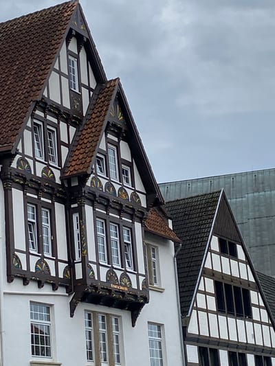 The image shows a close-up of the historic buildings in Bückeburg, Germany. The focus is on the detailed facades of two half-timbered houses, characterized by their white infill panels and dark wooden beams. These buildings feature steeply pitched, tiled roofs. Ornate decorative elements are visible on the eaves and window frames, with colorful patterns adding to their historical charm. Several windows with white frames are present, reflecting the overcast sky. In the background, a modern building with a metallic, ribbed facade is visible, creating a contrast between old and new architecture. The scene is captured on a cloudy day, with the sky occupying the upper portion of the frame. There are no people visible in the image.