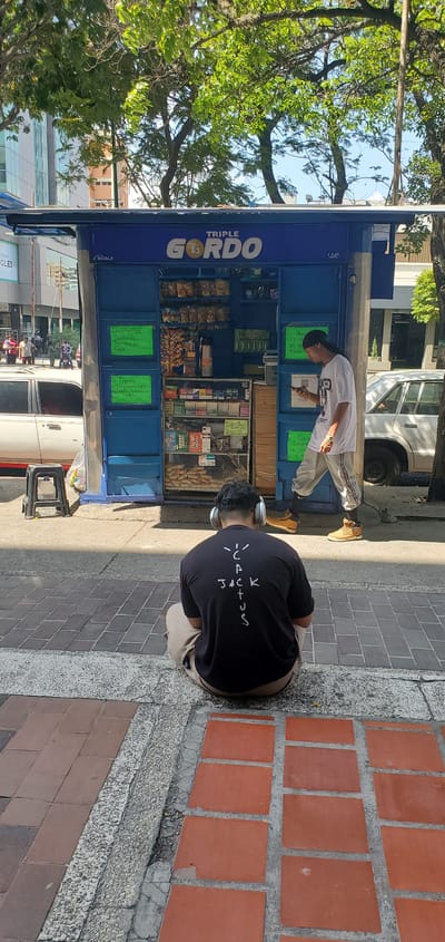 The scene captures a vibrant street-side setting in Barquisimeto, Venezuela. A "Triple Gordo" blue kiosk prominently displays the brand name on its facade. The kiosk features an open display of snacks and sweets. One person is standing in front of it looking at their phone. A second person is sitting on the sidewalk. The person is wearing a "Cactus Jack" T-shirt and headphones. The sidewalk is made of gray stone and red brick. A tree can be seen on the opposite side of the street. Several cars pass by. The buildings across the street appear to be modern. The setting suggests an urban, commercial area.