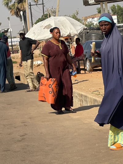 The scene depicts a daytime street environment in Basawa, Nigeria.

In the immediate foreground, a woman wearing a dark blue full-body hijab and a light blue headscarf beneath is positioned on the right side, looking directly towards the viewer. She holds a light-colored, elongated food item, possibly corn on the cob, in her right hand, with a thin strand of it visible extending from her mouth. Her visible lower garment is patterned light yellow and green, and she wears orange open-toed sandals.

Slightly to the left and behind her, a woman in a long, dark maroon short-sleeved dress with gold trim at the neckline and sleeve edges walks towards the left side of the frame. She carries a large, rectangular orange woven bag with black circular patterns in her left hand and a small, blue and orange object in her right. Her dark hair is pulled back.

In the mid-ground, a man wearing a black short-sleeved t-shirt, light brown trousers, and a baseball cap stands with his hands on his hips, facing right. A black backpack is on the paved ground near his feet, adjacent to a concrete drainage ditch. To the right of the man and behind the maroon-dressed woman, a large, white, somewhat torn canopy umbrella provides shade. Beneath it, a dark-colored sedan is parked, and a woman in a red long-sleeved shirt and patterned trousers is seated on a stool, facing left. Behind the parked car, several large black water storage tanks are visible.

The environment includes a paved road surface in the foreground, transitioning to a dirt shoulder on the right, separated by a concrete curb. Utility poles with overhead wires are visible in the background, along with sparse trees and partially obscured commercial signage, including a sign with "AGO". The sky is clear and light, indicating daylight.