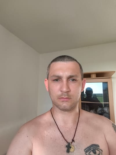 This is a medium shot, front-facing selfie of a shirtless man with a buzz cut and light stubble. He has light skin with some freckles and a few minor scars on his chest and shoulder. He is wearing a dark cord necklace with a cross and a circular pendant. In the background, there is a light-colored wall and a wooden cabinet with a glass door. Inside the cabinet, a reflection shows another person, presumably the subject, holding a phone, taking the picture. The reflection also shows a landscape with a field and sky, suggesting the cabinet is near a window. The lighting suggests it is daytime. The man's expression is neutral, with a slight frown. There is a tattoo on his chest, partially visible, which appears to be an eye with lines above it.