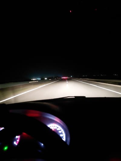 A nighttime perspective from inside a moving car. The illuminated dashboard displays a speedometer with red accents. A steering wheel is partially visible in the foreground. Outside, road markings and distant lights create a visual perspective. A highway lane is distinguished by white lines, leading to the glow of other vehicle lights. The scene is dark, highlighting the contrast between interior car lights and the external environment.