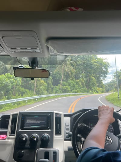 This media file depicts the interior of a vehicle, likely a Toyota van, as it is driven along a paved road. The perspective is from inside the vehicle, looking forward and slightly to the right. A person's right hand is visible on the steering wheel, indicating they are the driver. The steering wheel features a Toyota emblem and an instrument cluster showing speed and other vehicle information.
The dashboard includes a touchscreen infotainment system displaying a caution message about driving safety. Several buttons and knobs for climate control and other functions are also visible. The gear shifter is in the "R" position, suggesting the vehicle is in reverse or parked.
Outside the vehicle, through the windshield, a lush green landscape with trees and palm trees is visible. The road ahead curves to the right, bordered by a metal guardrail and grassy verges. The sky appears overcast, suggesting a cloudy or potentially rainy day.
The scene is set on a road in Ban Wang Pra Chan, Thailand. The overall impression is one of traveling through a tropical or subtropical environment. No specific emotions are evident, but the presence of a driver indicates an ongoing journey or activity. The visible text includes the infotainment system's caution message, Toyota branding, and labels for vehicle functions like "ROOM" and "REAR". There are also numerical readings on the instrument cluster.