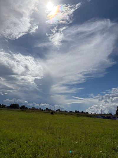 The image depicts a vast green field under a dramatic sky filled with a mix of white and gray clouds. The sun is high in the sky, partially obscured by clouds, and a subtle rainbow-like effect is visible near it. In the distance, a treeline marks the horizon, beyond which is a road with scattered streetlights and some structures, possibly a commercial area. The field itself is characterized by tall, lush green grass with some scattered wildflowers. The scene appears to be captured in an open, undeveloped area, likely on the outskirts of a populated area. The overall impression is one of natural beauty and open space. The specific location in Jēkabpils, Latvia, is an open field adjacent to a road, with a commercial zone visible in the background.