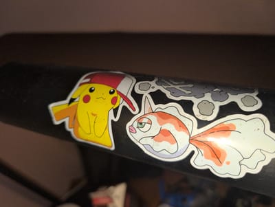 In an indoor, dimly lit environment, two stickers are adhered to a black, cylindrical object, possibly a baseball bat. The sticker on the left features a yellow cartoon character resembling Pikachu, wearing a red and white hat. To the right of Pikachu is a sticker of a koi fish with orange and white markings and a green eye. The background is blurred and appears to be a dark brown surface. The location is the city of Kingston, Canada, and it's not possible to determine any further details of the city or its surroundings from the image.