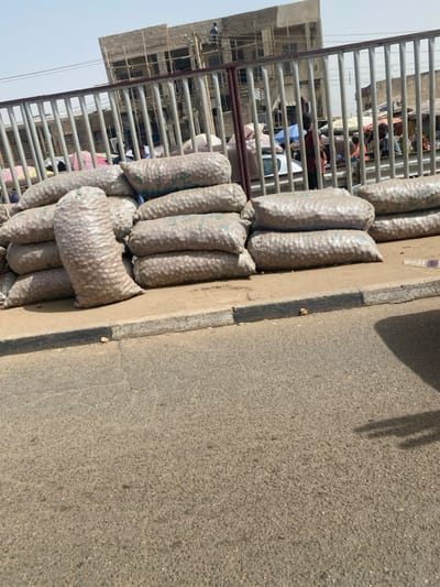 This image depicts a street scene in Rijiyar Lemo, Nigeria, characterized by commercial activity. In the foreground and midground, numerous large, clear plastic sacks filled with light brown, roughly spherical produce (likely potatoes or similar tubers) are stacked on a grey concrete sidewalk. These sacks form several piles, with one elongated sack standing vertically against a horizontal stack. The sidewalk features a curb painted black and white, separating it from a textured, grey asphalt road that occupies the lower half of the frame. A dark shadow, possibly from an unseen vehicle, extends into the lower right corner of the road.
Behind the stacked sacks, a light-colored metal fence with vertical bars runs horizontally across the image, serving as a barrier to a bustling background area. Beyond this fence, a market scene unfolds with several individuals. One person wearing a red shirt is visible standing near the fence. Other individuals are partially obscured by market stalls and tarpaulin shelters, some of which are blue. A white vehicle is partially visible among the market activity. In the far background, a multi-story building under construction, featuring an unfinished concrete structure and visible scaffolding, dominates the left side. A lone individual, identifiable by a blue top, is positioned on the upper level or roof of this building, seemingly engaged in work. The sky overhead is a hazy, light blue.