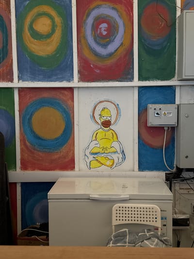 The image shows a brightly painted wall with concentric circles and a depiction of Homer Simpson in a meditating pose, adorned with a halo and pearls. To the right of the painting, a gray electrical box is mounted on the wall, with wires running from it. Below the wall, a white chest freezer is visible, with a small, white plastic chair in front of it. The chair has a checkered blanket draped over it. The scene appears to be in an outdoor or semi-outdoor setting, possibly a shop or public space, given the presence of the freezer and electrical equipment. The style of the artwork suggests a whimsical or humorous artistic expression. The overall impression is of a slightly cluttered but visually interesting environment. The image does not provide explicit information about the location in Koshkol', Kyrgyzstan, beyond its presence within the city.