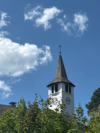 A clear blue sky with scattered white clouds is visible above a church tower in Titisee-Neustadt, Germany. The church tower is white with a dark, conical roof, topped with a cross. Two clock faces are visible on the tower, one showing approximately 3:05 and the other around 12:10. Green trees and the dark-tiled roof of a building partially obscure the base of the tower. Several small, pink dots are scattered across the sky in the upper left quadrant of the image.
