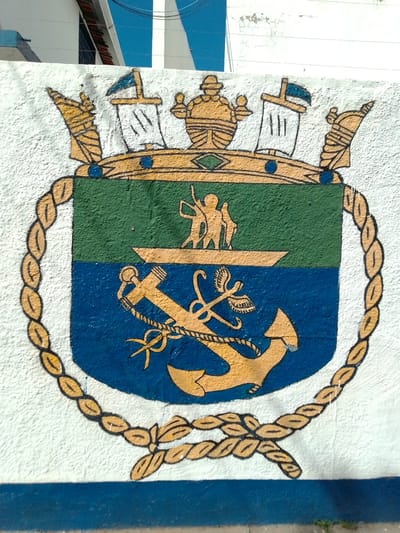 This is a close-up, eye-level outdoor photograph of a mural painted on a textured, white stucco wall. The mural depicts a coat of arms, likely with maritime or military significance. The coat of arms is framed by a thick, braided yellow rope. The shield itself is divided horizontally into two sections: a green upper section and a blue lower section. The upper green section features a yellow boat with two small figures standing in it. The lower blue section contains a large, prominent yellow anchor with a rope wrapped around its shank and fluke. Above the shield, a golden crown-like structure is depicted with various decorative elements, including flags on masts and what appear to be figures or statues. The colors used are primarily green, blue, and yellow, with black outlines defining the shapes. The wall shows signs of wear and tear, with some chipping and cracking in the paint and stucco. In the background, a portion of a building with white and blue architectural elements is visible, as well as a clear blue sky. The mural is located on an exterior wall, possibly in the city of Cáceres, Brazil, based on the context provided.
