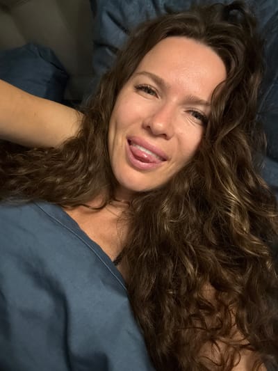 In an indoor setting, likely a bedroom with blue bedding, a woman with long, wavy brown hair is lying down and looking at the camera. She is wearing a blue garment, possibly a top or dress. Her right hand is visible behind her head, and she is sticking her tongue out playfully, winking with her right eye. The lighting suggests it is either nighttime or a dimly lit room. There is no visible indication of the city of Изумруд Снт, Russia, in the image.