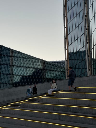 The image depicts a group of people on a wide, outdoor staircase illuminated by yellow lights embedded in the steps. The staircase is adjacent to a modern, glass-facade skyscraper. The building's reflective surface shows the sky and some of its own architecture. The overall scene suggests an urban environment, possibly a public space designed with contemporary architectural elements.

There are at least three individuals visible on the stairs. One person, wearing a dark hoodie and blue jeans, appears to be standing on a higher step, with their back to the camera. Further down the stairs, two people are more clearly visible. One, a woman wearing a light-colored jacket and white pants, is looking towards the camera. Another person, wearing a dark jacket, is standing next to her, also facing somewhat towards the camera. A fourth person, wearing a blue denim jacket and a hijab, is visible on a lower step, looking down.

The lighting suggests it might be evening or dusk, as the yellow lights on the stairs are prominent against the backdrop of the darkening sky and the building's facade. The weather appears to be clear, with a bright, pale sky. The architecture of the skyscraper, with its geometric pattern of glass and metal, is a significant feature of the scene. The image is oriented sideways, with the skyscraper and the sky occupying the left side and the staircase with people on the right.