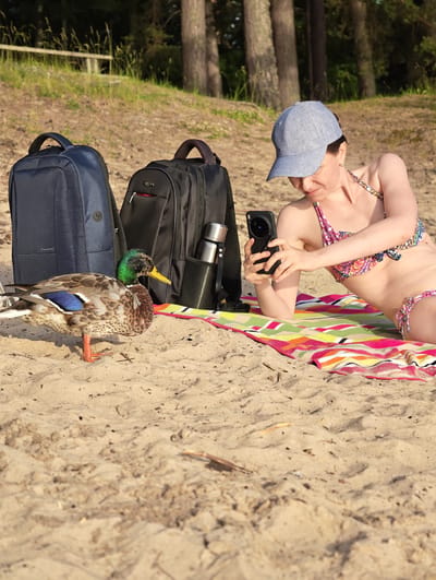 A woman wearing a bikini and a baseball cap is lying on a striped towel on a sandy beach, taking a picture with her phone. Two backpacks, a water bottle, and a duck are also on the beach near her. The scene appears to be set in Motala, Sweden, during the daytime, likely in the summer due to the beach setting and the woman's attire. The weather seems pleasant, with sunlight casting shadows on the sand. The woman's focus on her phone suggests she is capturing a moment or interacting with her device. The presence of the duck adds a natural element to the scene, and its proximity to the woman could indicate it is accustomed to human presence or was attracted by something. The background features trees and a wooden fence, suggesting a natural or park-like area adjacent to the beach. The overall mood of the image is relaxed and recreational. There is no visible text in the image.