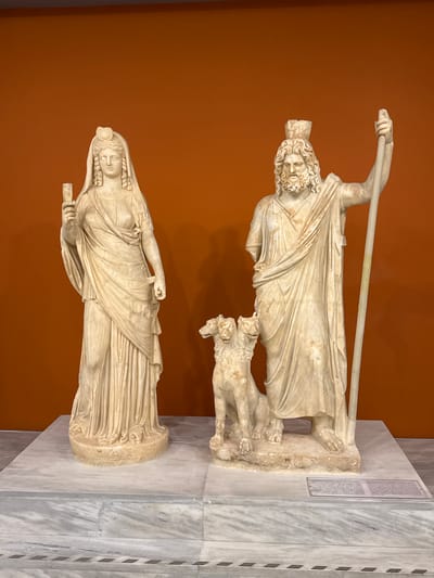 This image shows two marble statues in a museum setting. The statue on the left depicts a female figure wearing a veil and holding a rectangular object in her raised left hand. She is draped in flowing robes. The statue on the right depicts a male figure with a beard and a crown-like headdress, holding a staff in his right hand and standing next to a three-headed dog. He is also draped in flowing robes. Both statues are standing on separate marble bases, which are placed on a larger marble platform. The background is a solid, orange-brown color. A plaque with text is visible on the platform in front of the male statue. This is a photograph of an exhibit at the Heraklion Archaeological Museum in Heraklion, Greece, likely depicting figures from Greek mythology.