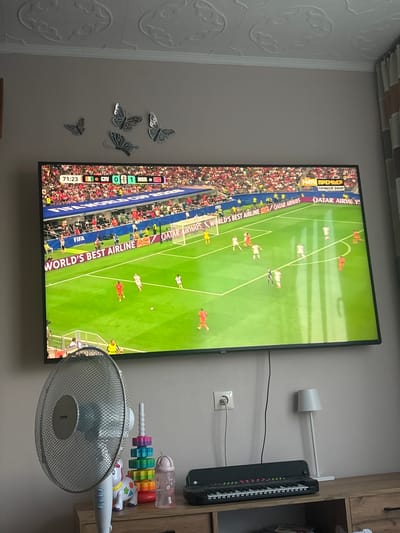 In a room with beige walls and a textured ceiling, a large flat-screen television displays a soccer match. The scoreboard indicates the time is 71:23, with one team, "CIV," scoring 0 and the other, "NOR," scoring 1. The game is part of the "FIFA WORLD CUP 2020." Several decorative butterflies are affixed to the wall above the television. Below the TV, a floor fan with a metallic grille is positioned to the left, while a small keyboard and a stack of colorful toys are on a wooden cabinet to the right. A white lamp stands next to the cabinet. The overall scene suggests a home environment where a sporting event is being watched. The location is identified as Krāslava, Latvia.