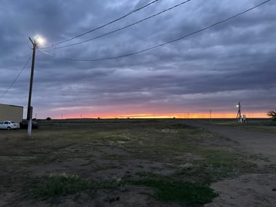 The image depicts a rural landscape at dusk in Өндіріс, Kazakhstan. The sky is filled with dramatic, dark clouds, hinting at a recent or approaching storm, though a band of warm, orange and pink light along the horizon suggests the sun is setting. Several power lines stretch across the frame, supported by a wooden pole with a bright light illuminating the foreground. In the middle ground, a dirt road winds through a sparsely vegetated field. Two cars are parked near a building on the left side of the frame. Further in the distance, telephone poles and a power transformer are visible. The overall mood is one of quiet solitude, transitioning from day to night under an atmospheric sky. There is no visible text in the image.